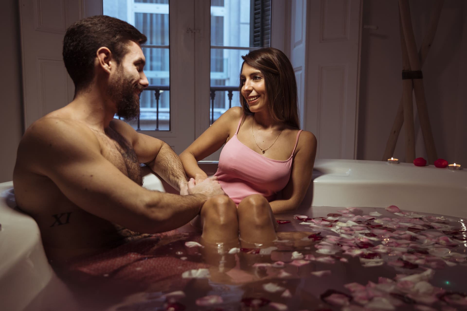 Casal sorridente aproveitando um banho relaxante em uma banheira com pétalas de rosa, criando um clima romântico e acolhedor.